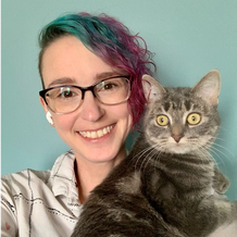 White woman with a full undercut dyed pink and blue wearing glasses and holding a grey cat with green eyes.